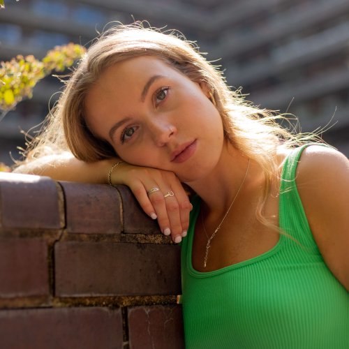 Graduate Eva Gadd wearing a bright green tank top and leaning against a red brick wall, with large block of flats in the background.