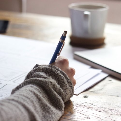 A hand holding a pen writing on paper with a mug in the background